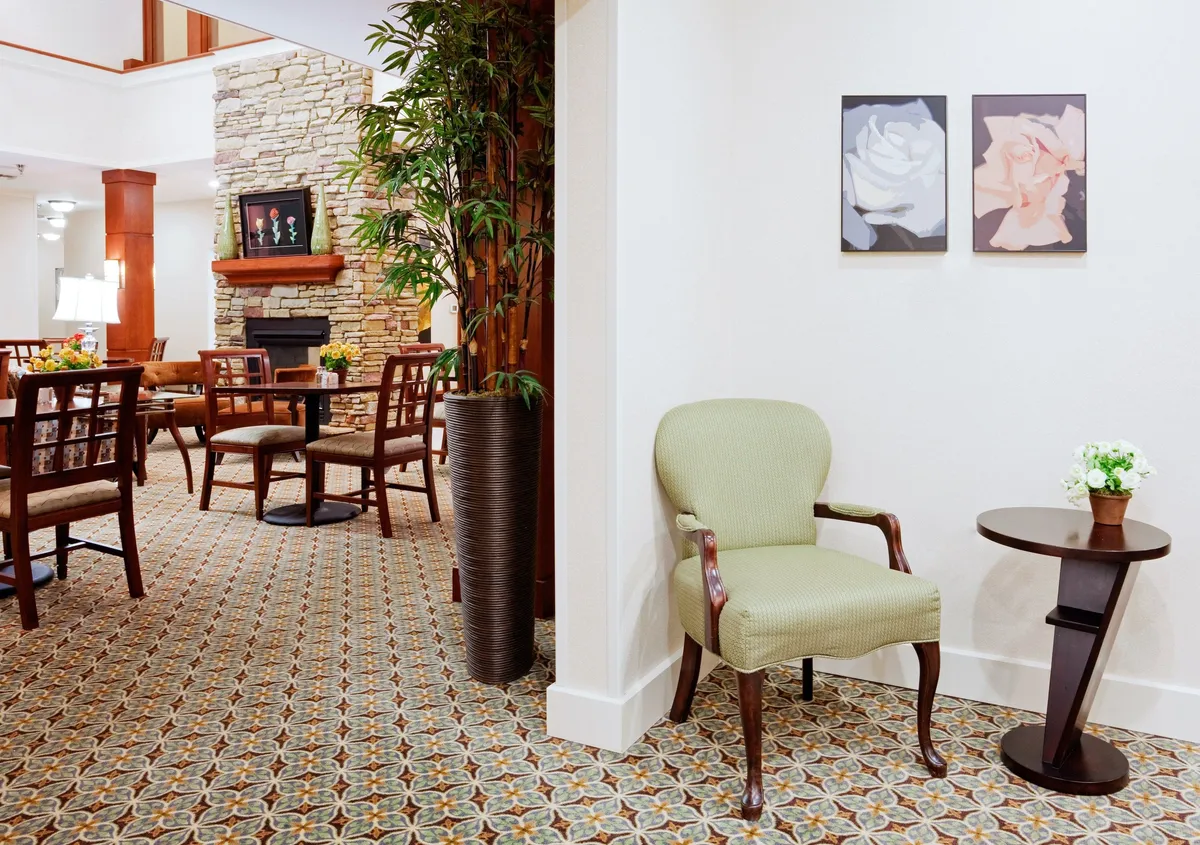 A welcoming and serene studio interior featuring a comfortable green chair, a small side table, floral wall art, and an indoor plant. In the background, a dining area invites guests to share meals or spend quiet moments together, all set in a space that feels both private and cozy.