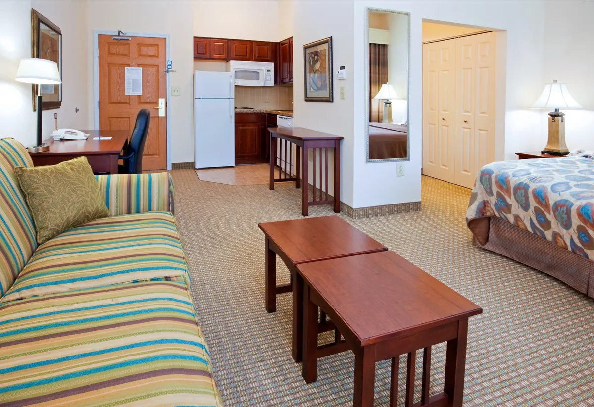A comfortable studio interior featuring a striped sofa, wooden tables, a visible kitchen area, and a bed with a decorative cover. The scene highlights the space's comfort and flexibility, with areas for relaxing, eating, and sleeping, all in a cohesive open-plan design.