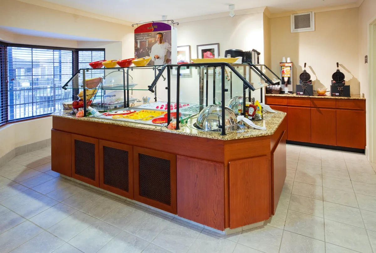 A vibrant breakfast buffet area featuring a well-stocked counter with colorful food displays, glass display cases filled with breakfast options, and multiple coffee machines. The scene is bright and welcoming, suggesting a space where guests gather in the morning to start their day.