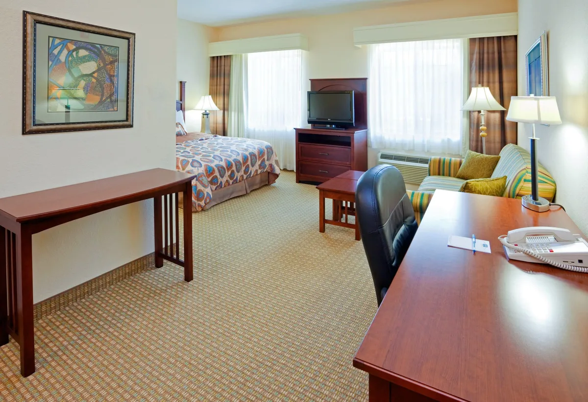 This image showcases a bright and welcoming studio space with a plush queen bed, a dedicated desk for work, a cozy sofa, flat-screen TV, and tasteful artwork. The arrangement blends comfort with practicality, highlighting the multi-functional nature of the suite. The furnishings and decor are individual, reflecting the uniqueness of each unit.